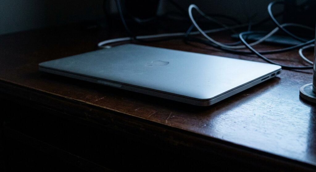 A MacBook Pro sitting closed on a dark desk at night, lit by a faint blue glow
